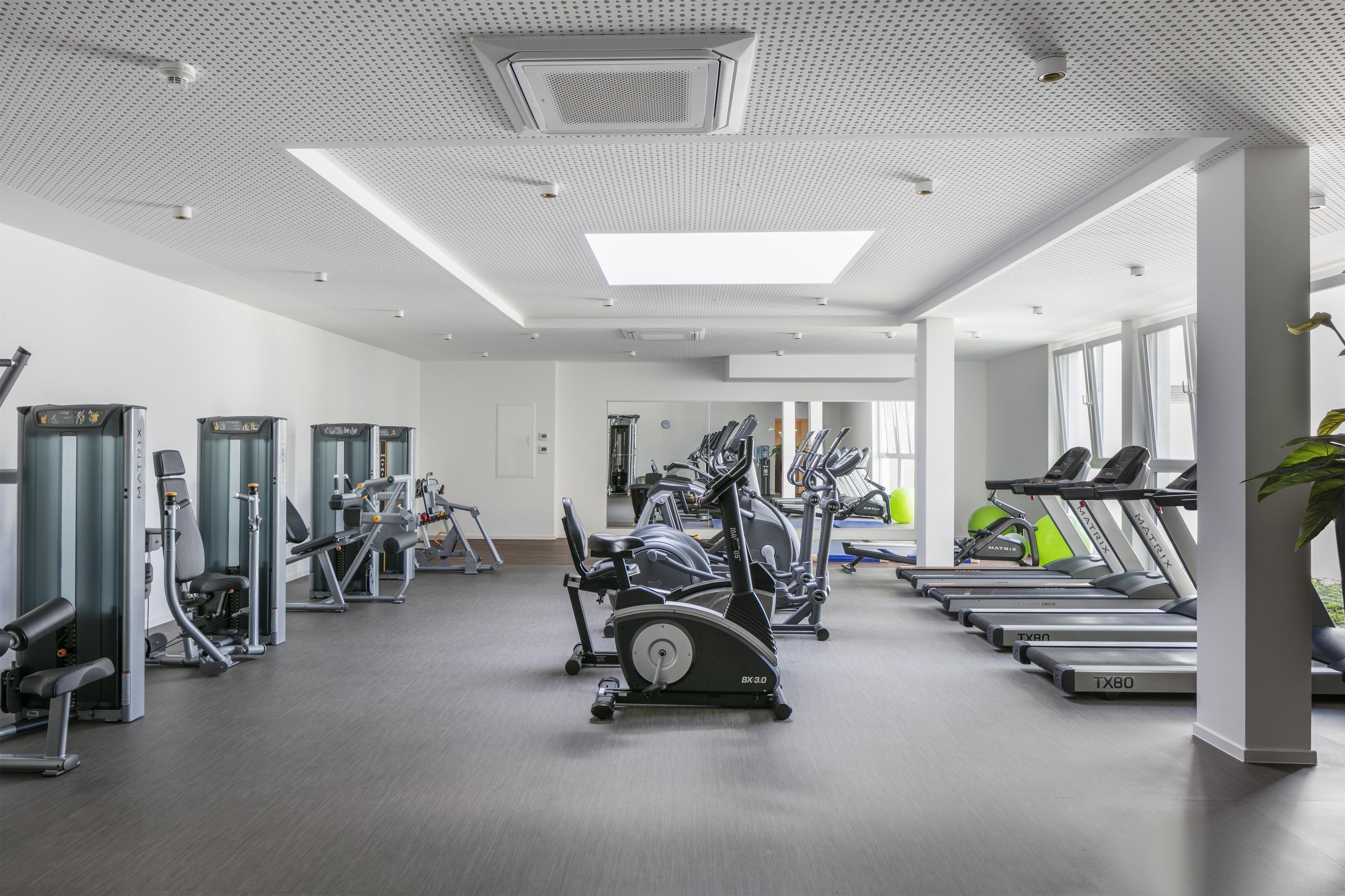 A gym with various exercise equipment, including treadmills and weight machines, and a large window providing natural light.
