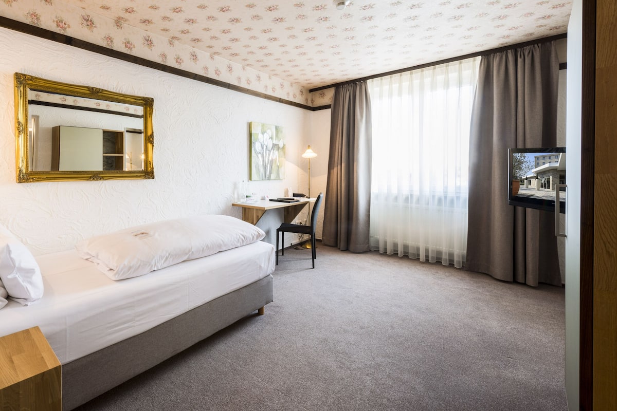 A hotel room with a bed, a desk, a chair, a TV, and a window with curtains.