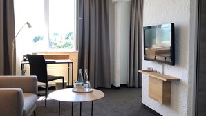 Ein Hotelzimmer mit einer Couch, einem kleinen runden Tisch mit Flaschen, einem Schreibtisch mit Stuhl, einem an der Wand befestigten Fernseher und einem großen Fenster mit Vorhängen.