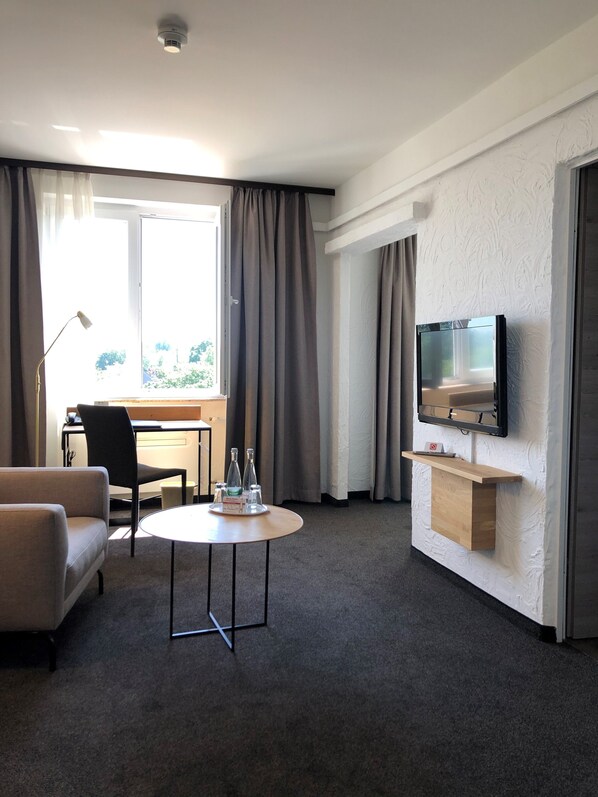 A hotel room with a sofa, a small round table with bottles, a desk with a chair, a TV mounted on the wall, and a large window with curtains.