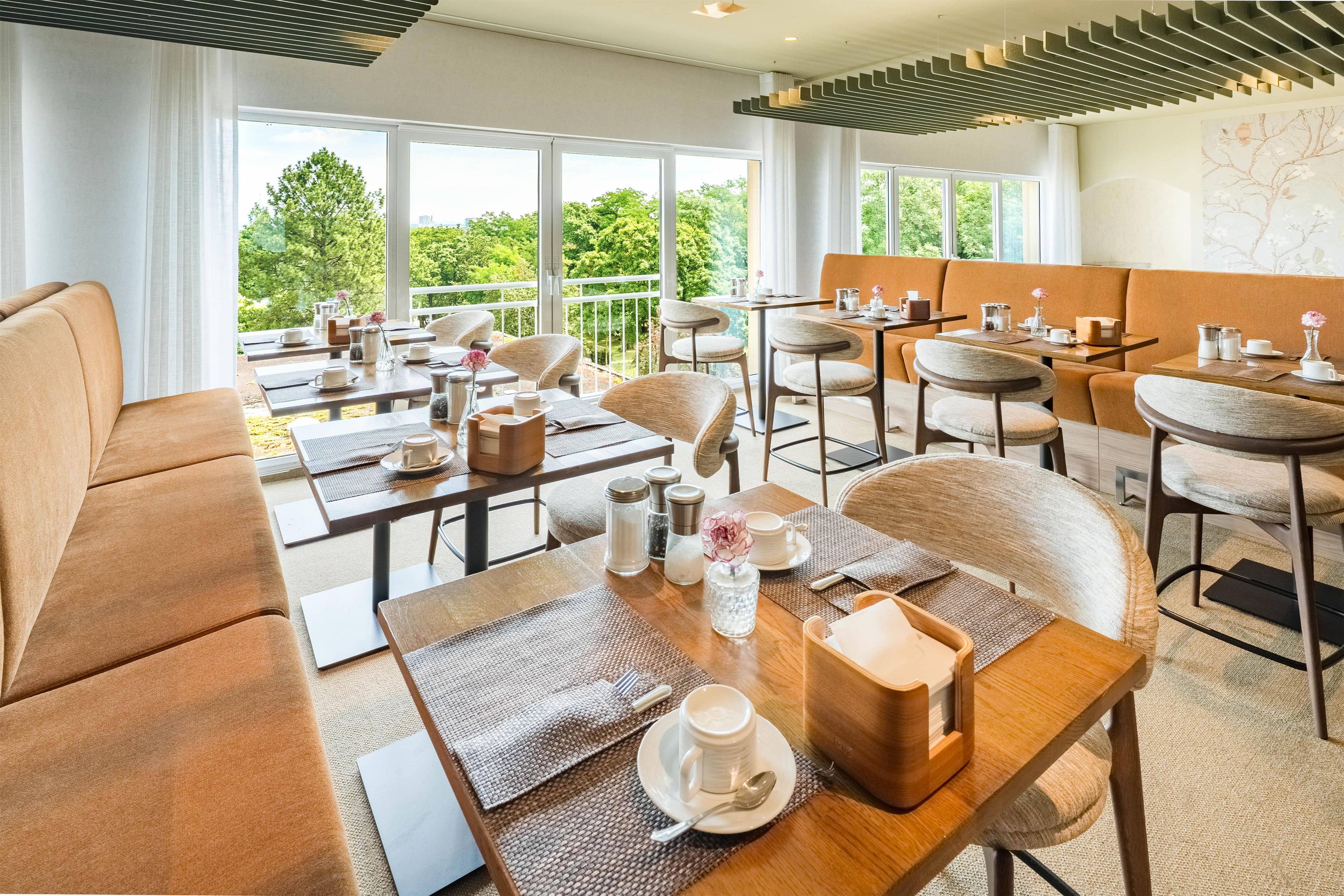 A modern dining area with multiple tables and chairs, large windows offering a view of greenery, and a contemporary ceiling design.