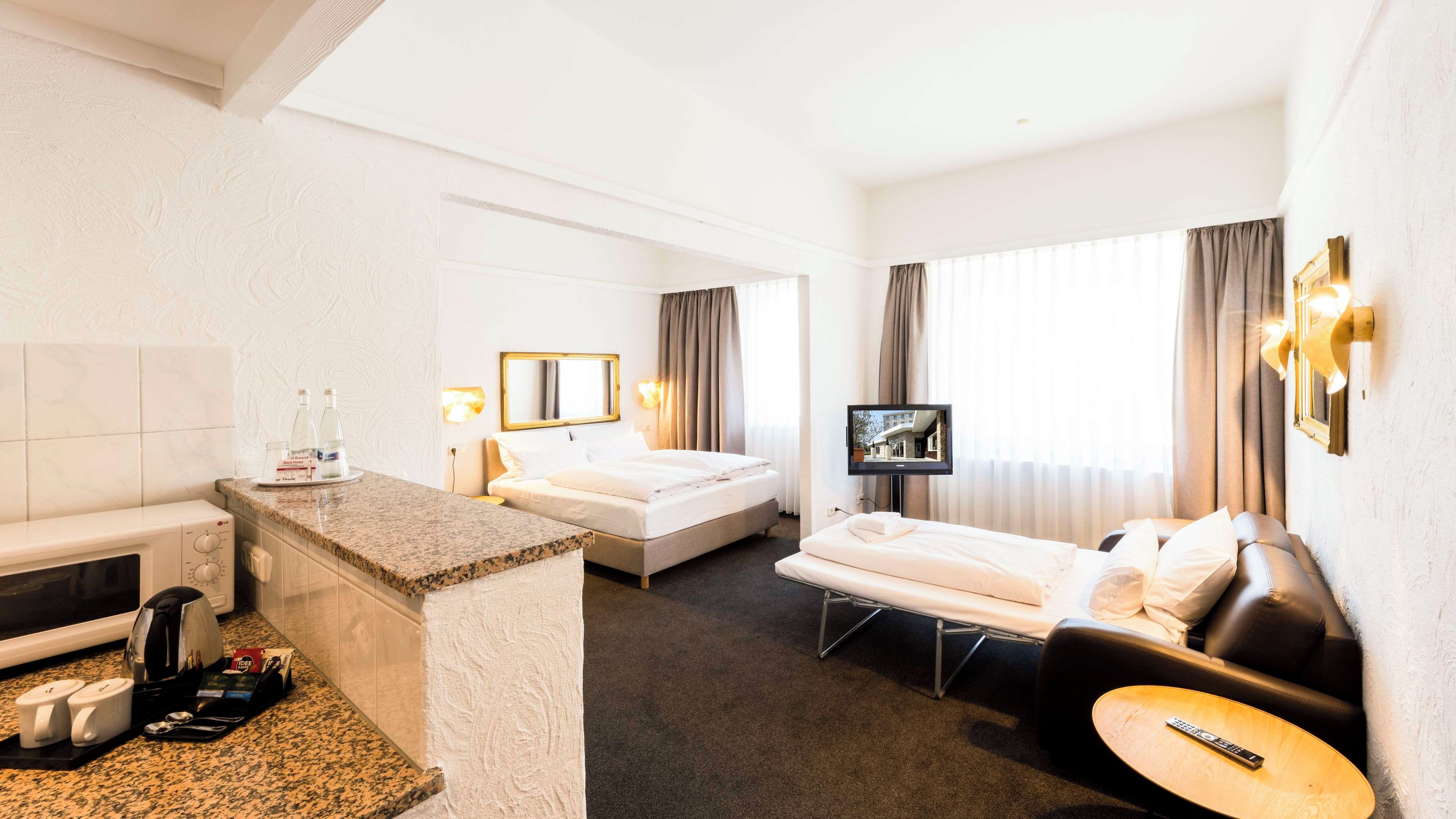 Una habitación de hotel con cama, sofá, televisión, pequeña mesa, microondas, cafetera y minibar.