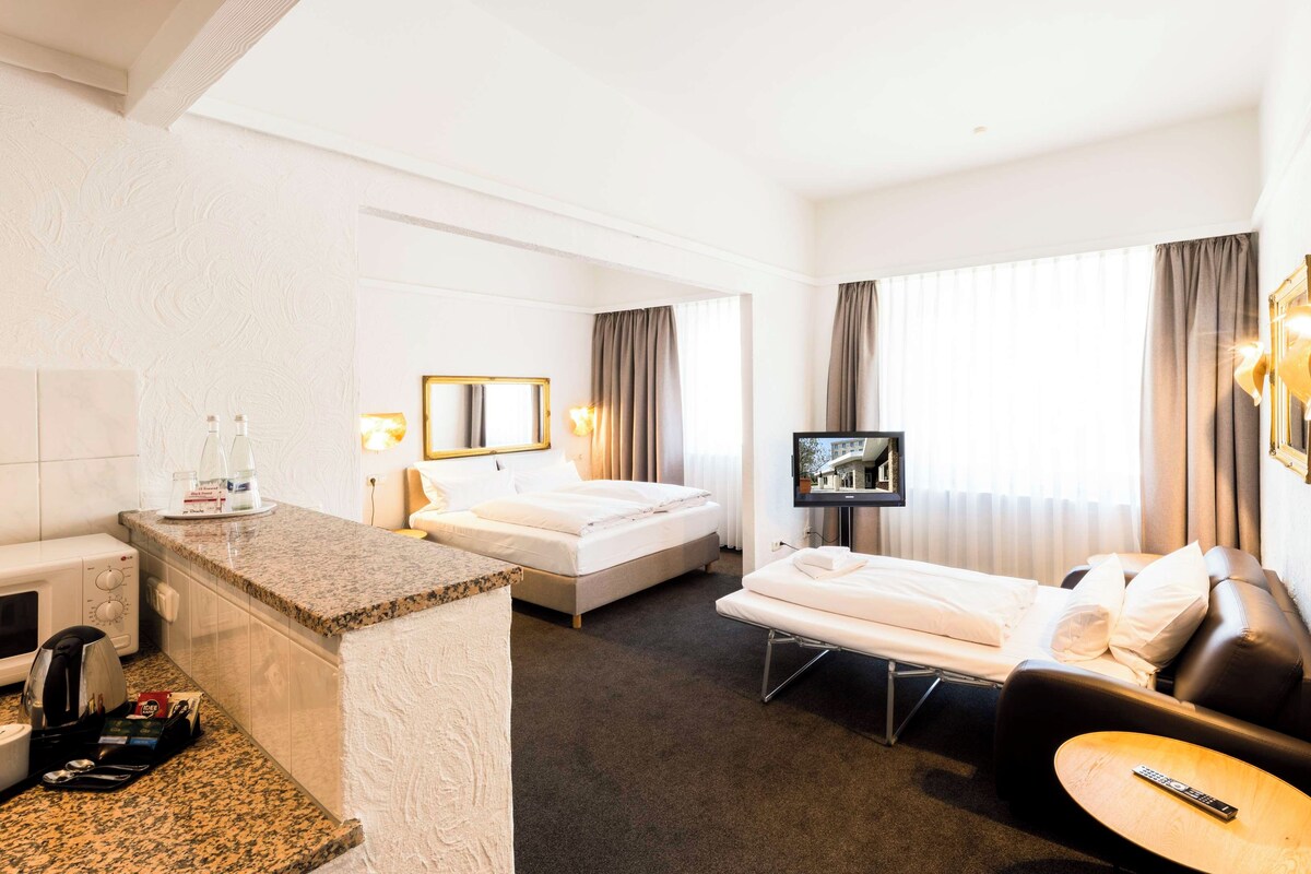 A hotel room with a bed, a sofa, a TV, a small table, a microwave, a coffee maker, and a minibar.