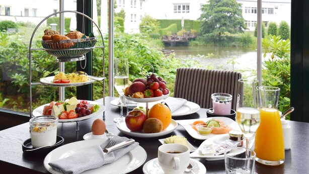 A breakfast setting with a variety of foods and beverages, a view of greenery and a building through a window. - Court Hotel (Halle Westphalia)