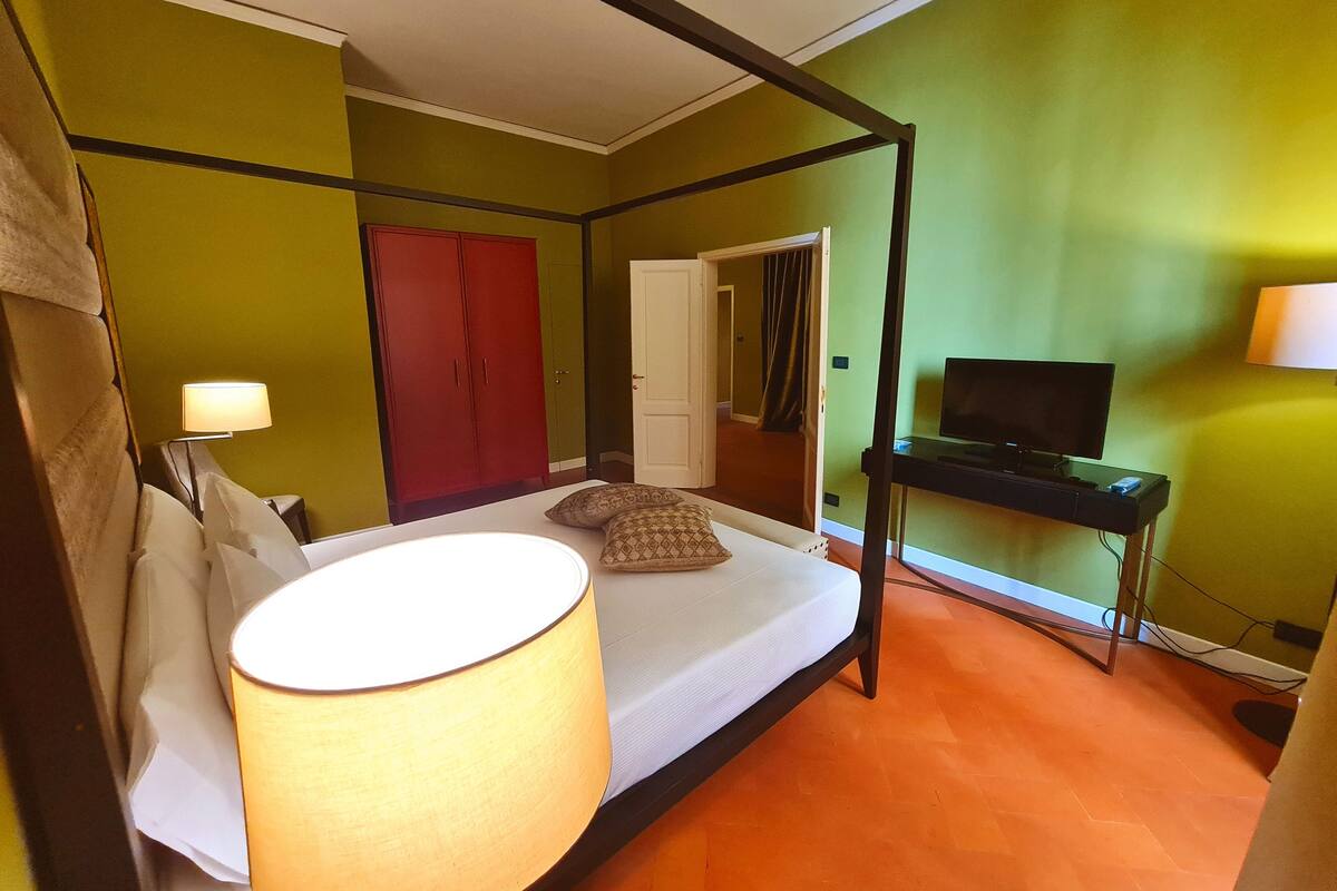 A room with a four-poster bed, a television on a stand, a red wardrobe, and a door leading to another room.