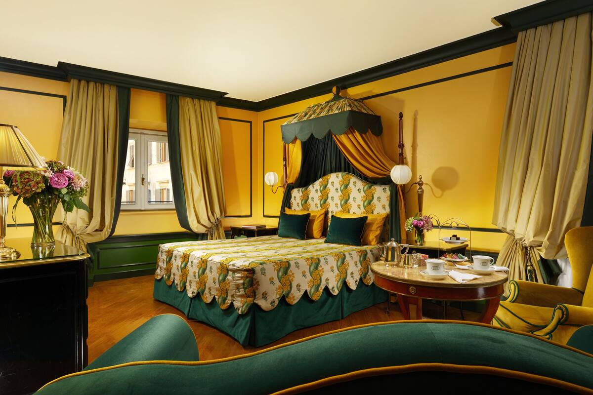 A hotel room with a bed, a round table with tea service, a chair, and a desk with a lamp and flowers.