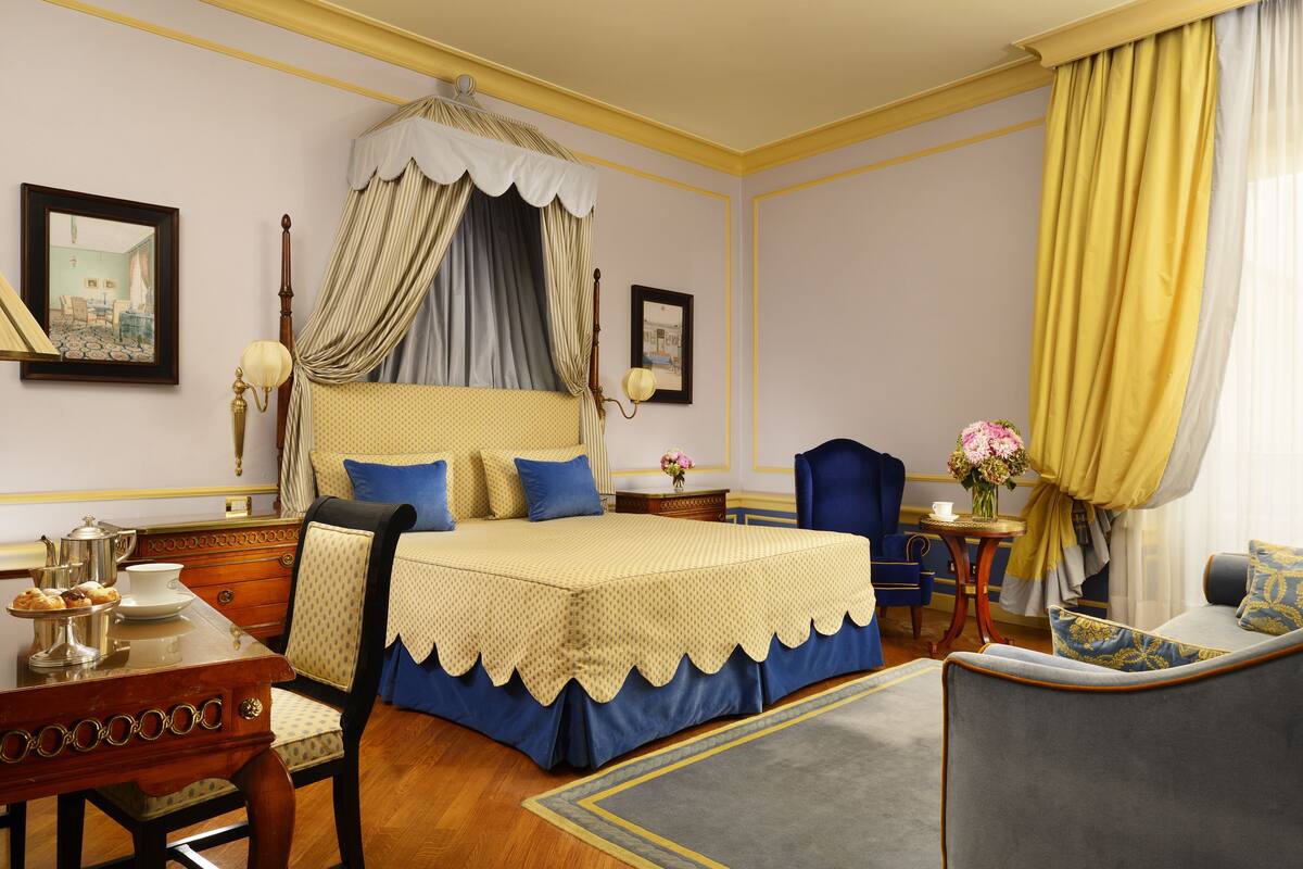 A bedroom with a four-poster bed, a desk with a chair, a small table with a vase of flowers, and a sofa.