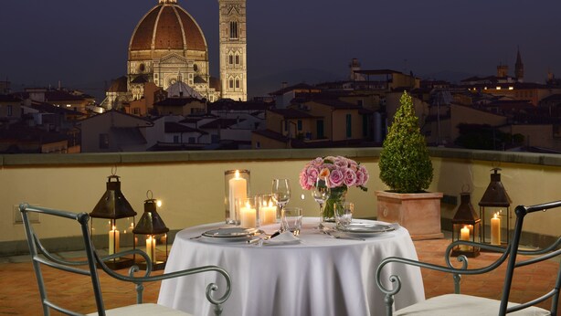 Terraza en la azotea con mesa lista para cenar, velas y un jarrón de flores, con vistas a un paisaje urbano histórico y arquitectura destacada.