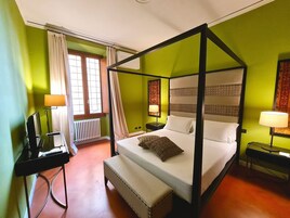A four-poster bed with a bench, a desk, and a chair in a room with green walls and a window with curtains.