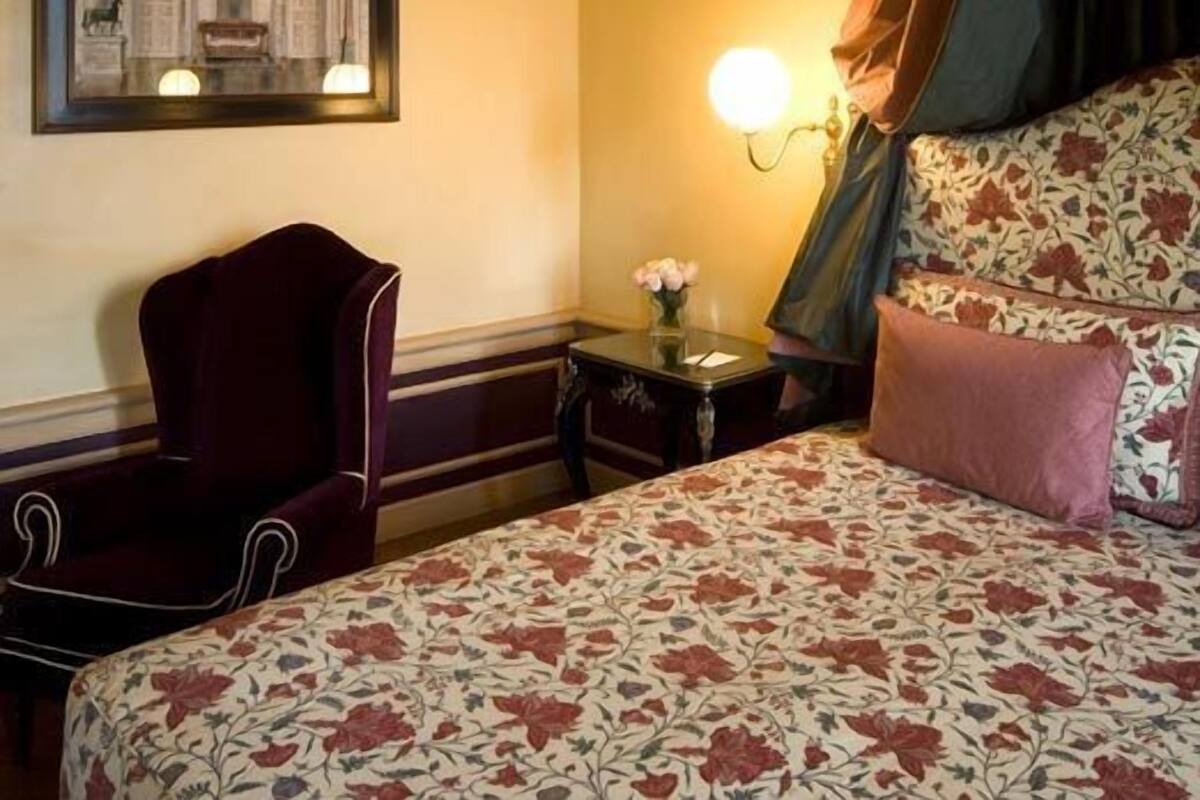 A hotel room with a bed, a chair, a nightstand with a lamp, and a framed picture on the wall.