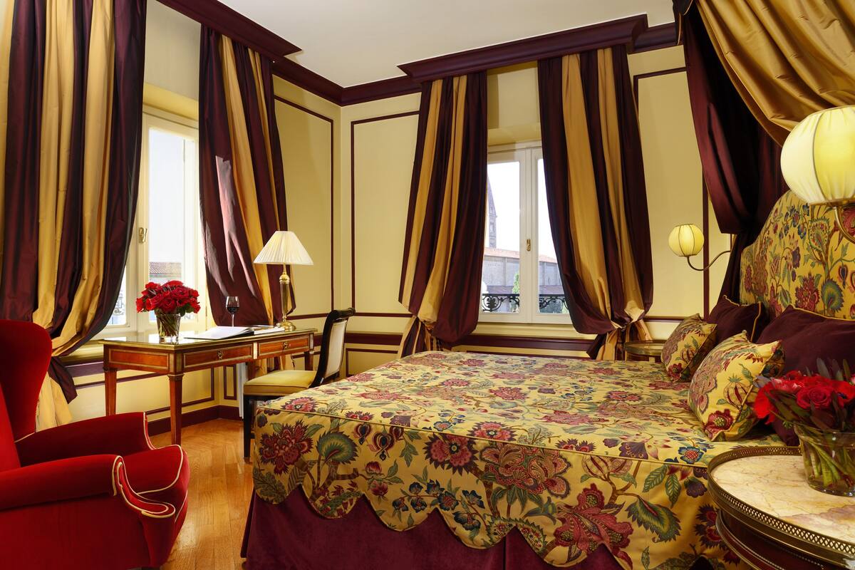 A hotel room with a large bed, a desk, a chair, and a red armchair. There are curtains on the windows and a vase with flowers on the desk.