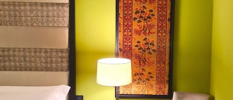 A hotel room with a bed, a chair, a table with a lamp, and a decorative tapestry on the wall.