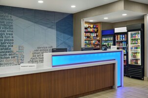 Zona de recepción de un hotel con mostrador, refrigerador lleno de bebidas y una pared con diseño geométrico.