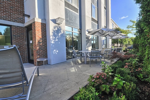 An outdoor patio with tables and chairs, surrounded by greenery and a building with large windows. - SpringHill Suites Detroit Southfield (Southfield)
