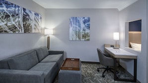 Una habitación de hotel moderna con un sofá gris, una mesita de madera, un escritorio con una silla y dos lámparas fijadas a la pared.