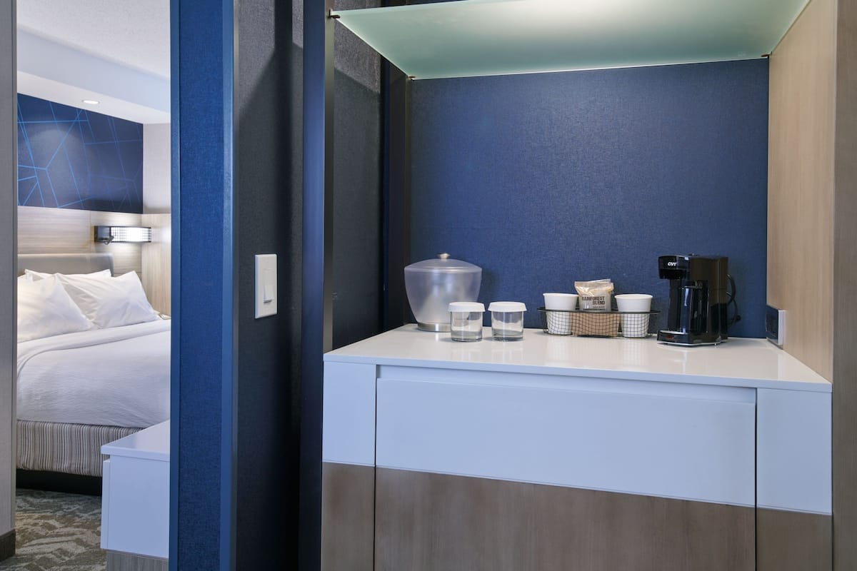 A hotel room with a bed, a small kitchenette with a coffee maker, and a breakfast bar with cups and a container.