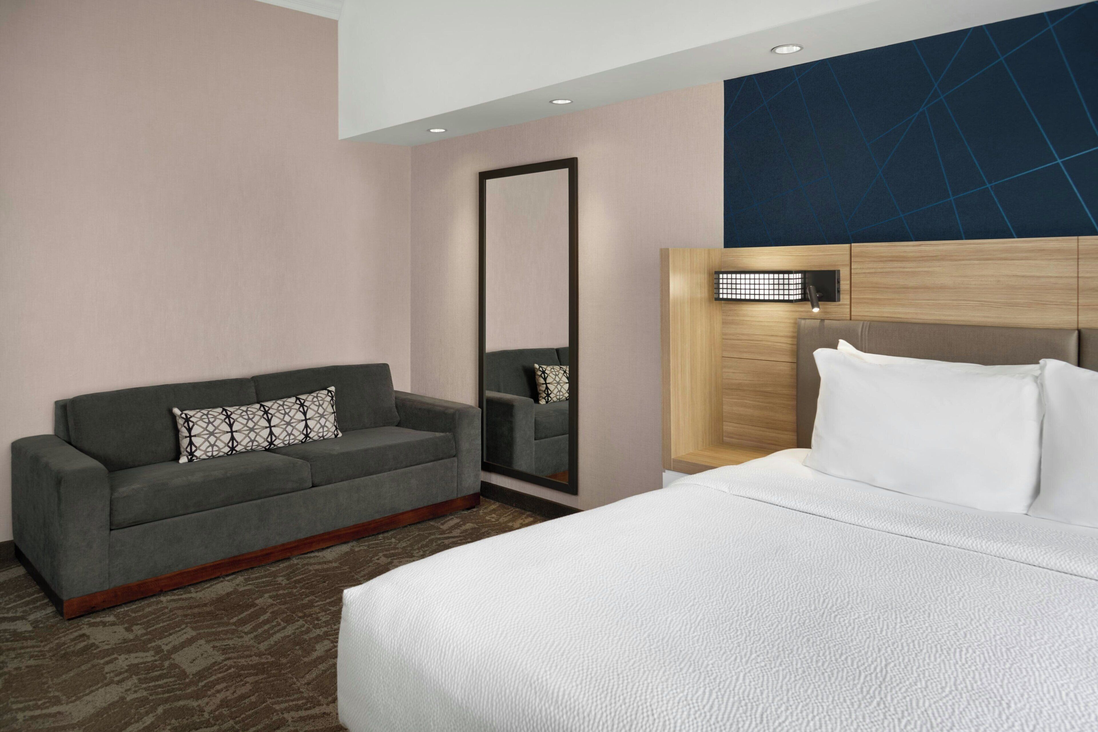 A hotel room with a bed, a sofa, a mirror, and a wall-mounted light fixture.