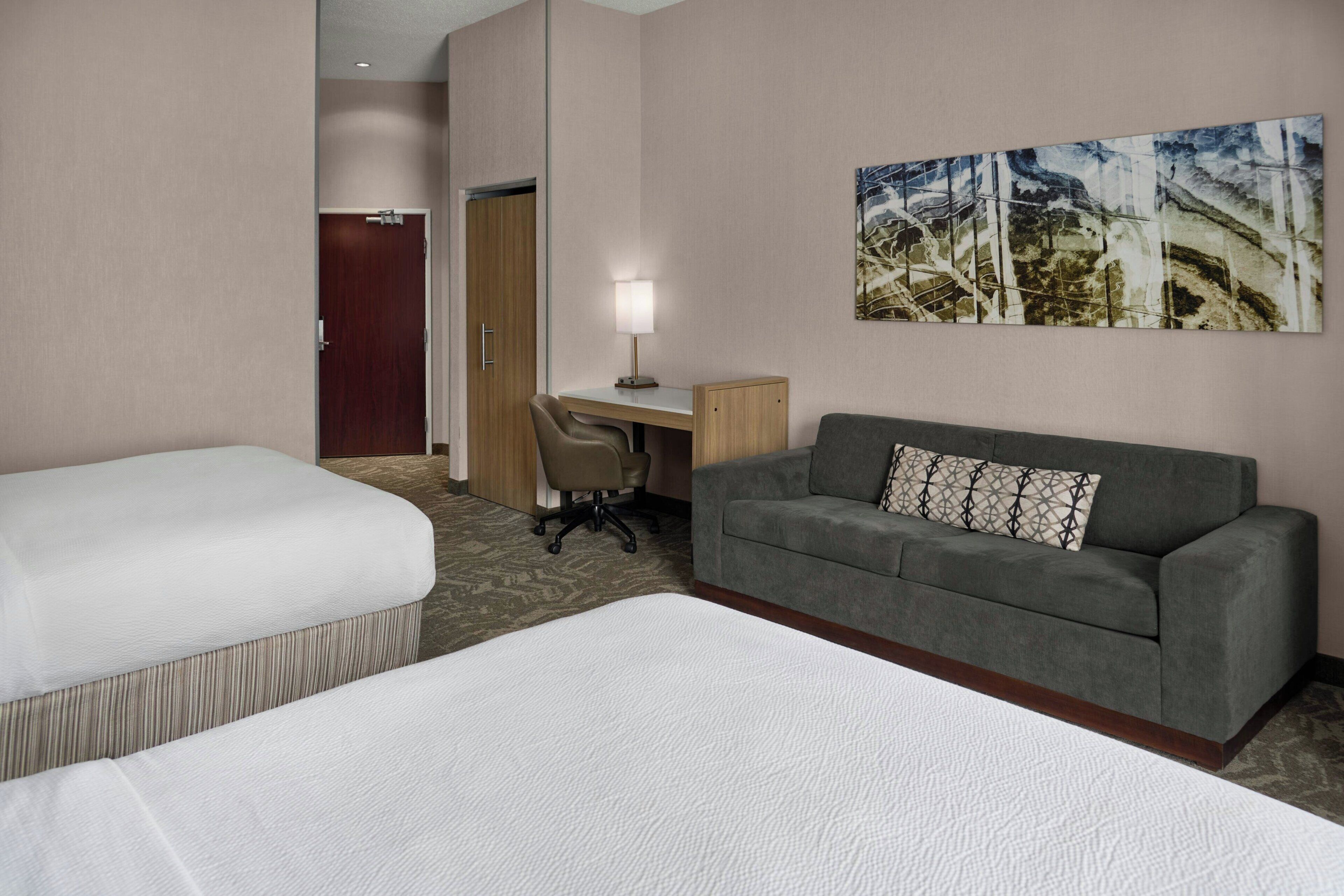 Habitación de hotel con dos camas individuales, un sofá gris con un cojín estampado, un escritorio con lámpara y un cuadro en la pared.