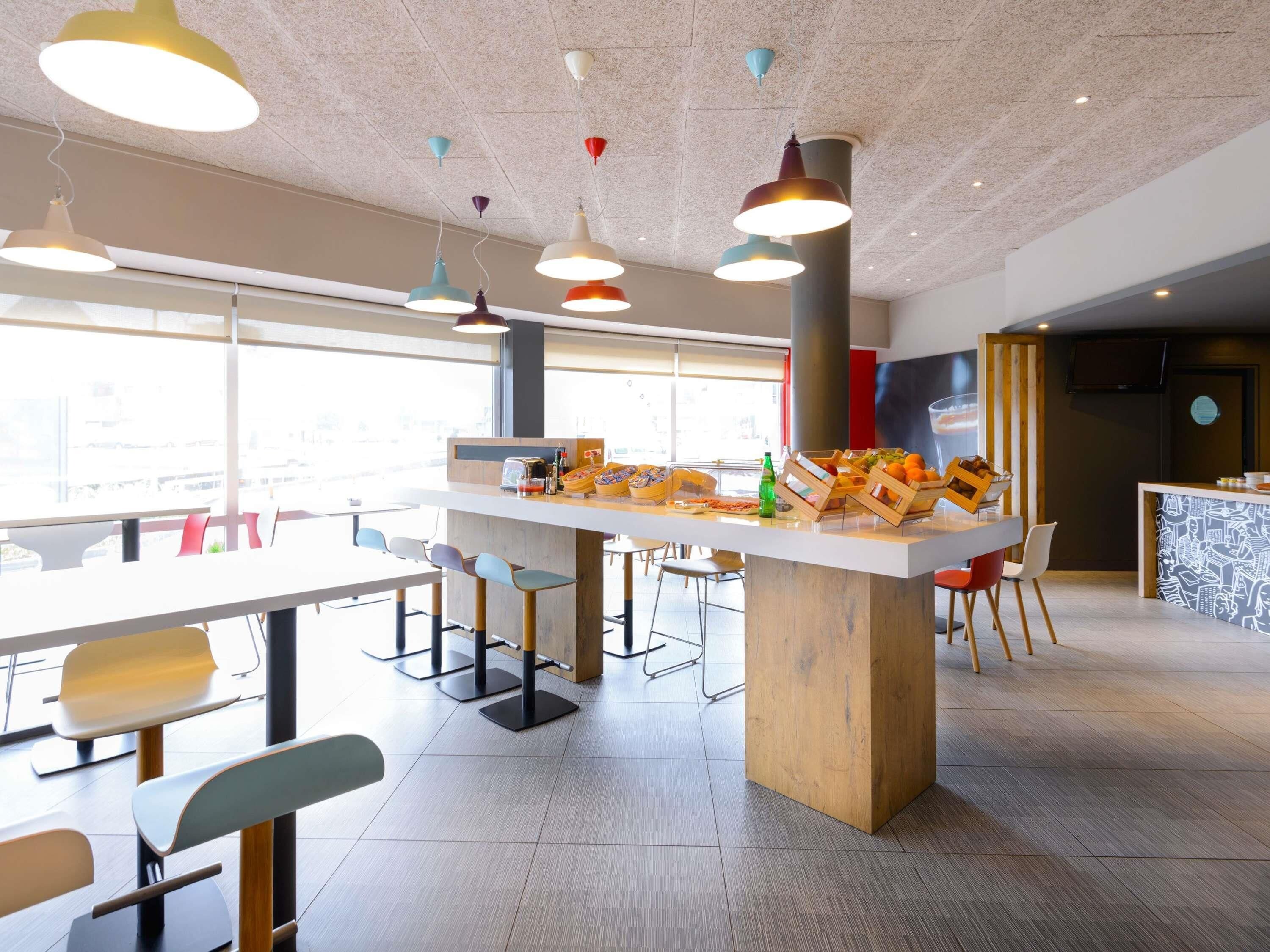 A modern cafe with a central wooden island, multiple seating options, and colorful pendant lighting.