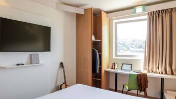 A bedroom with a bed, a TV, a guitar, a desk with a laptop, and a window with curtains.