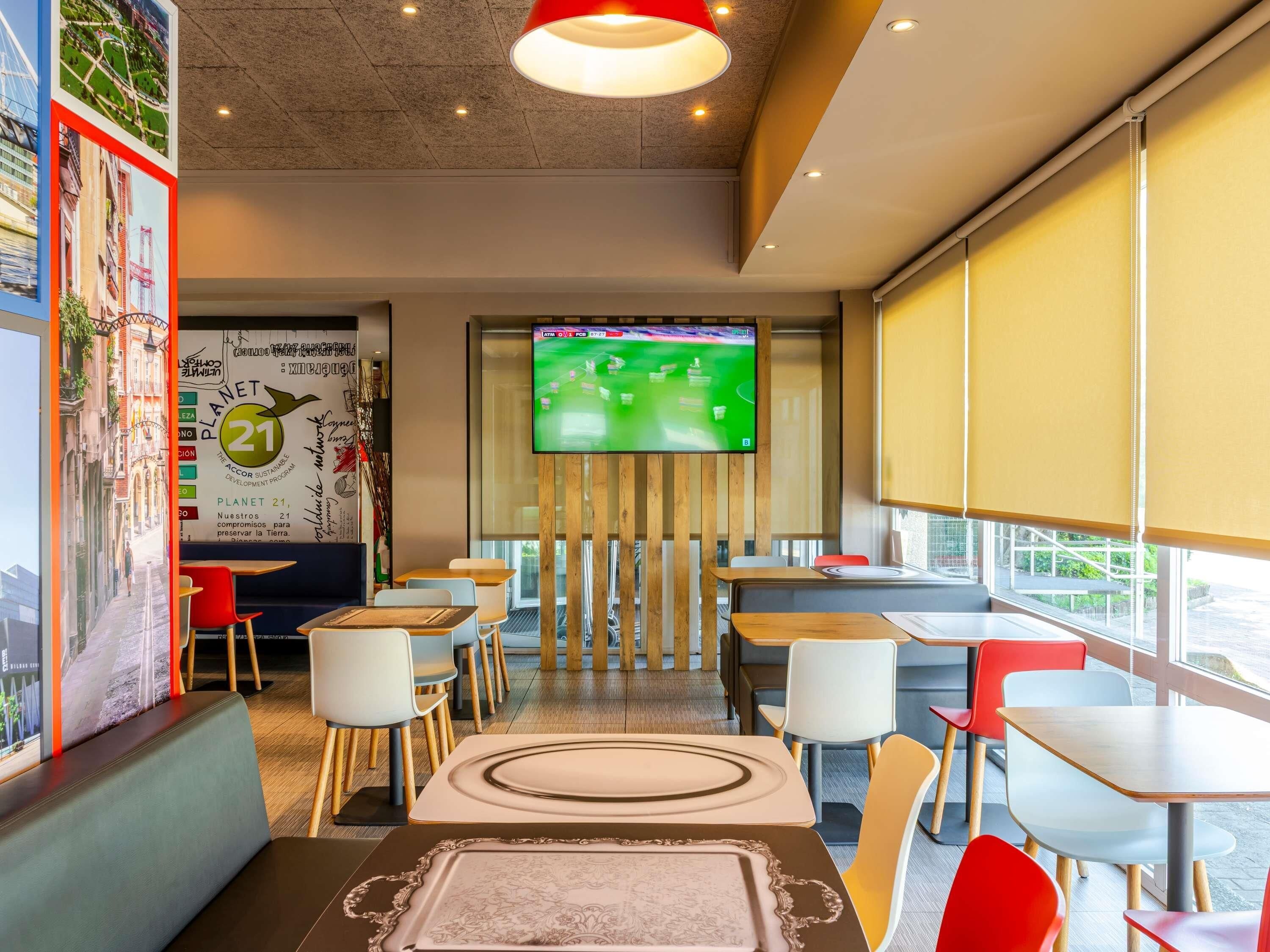 A modern restaurant with a large TV screen displaying a cricket match, wooden tables and chairs, and a wall with a 'Planet 21' mural.