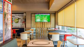 A modern restaurant with a large TV screen displaying a cricket match, wooden tables and chairs, and a wall with a 'Planet 21' mural.
