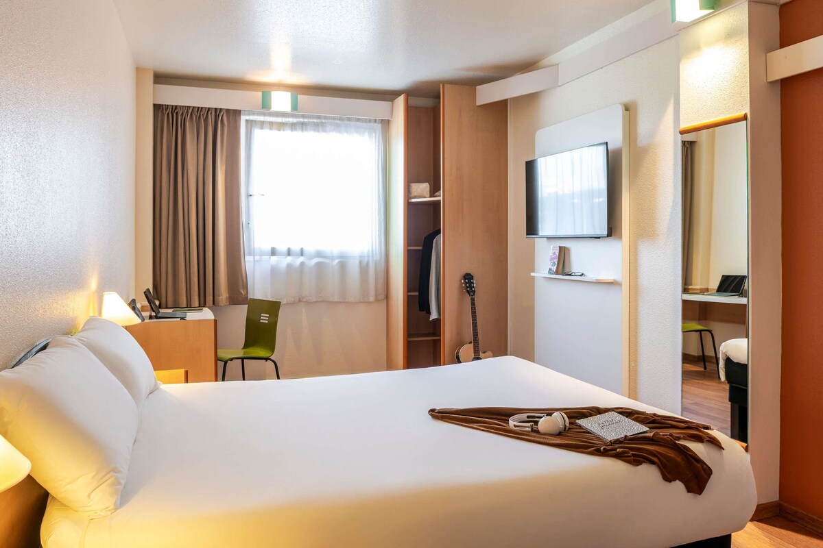 A hotel room with a large bed, a desk with a chair, a television, and a guitar.