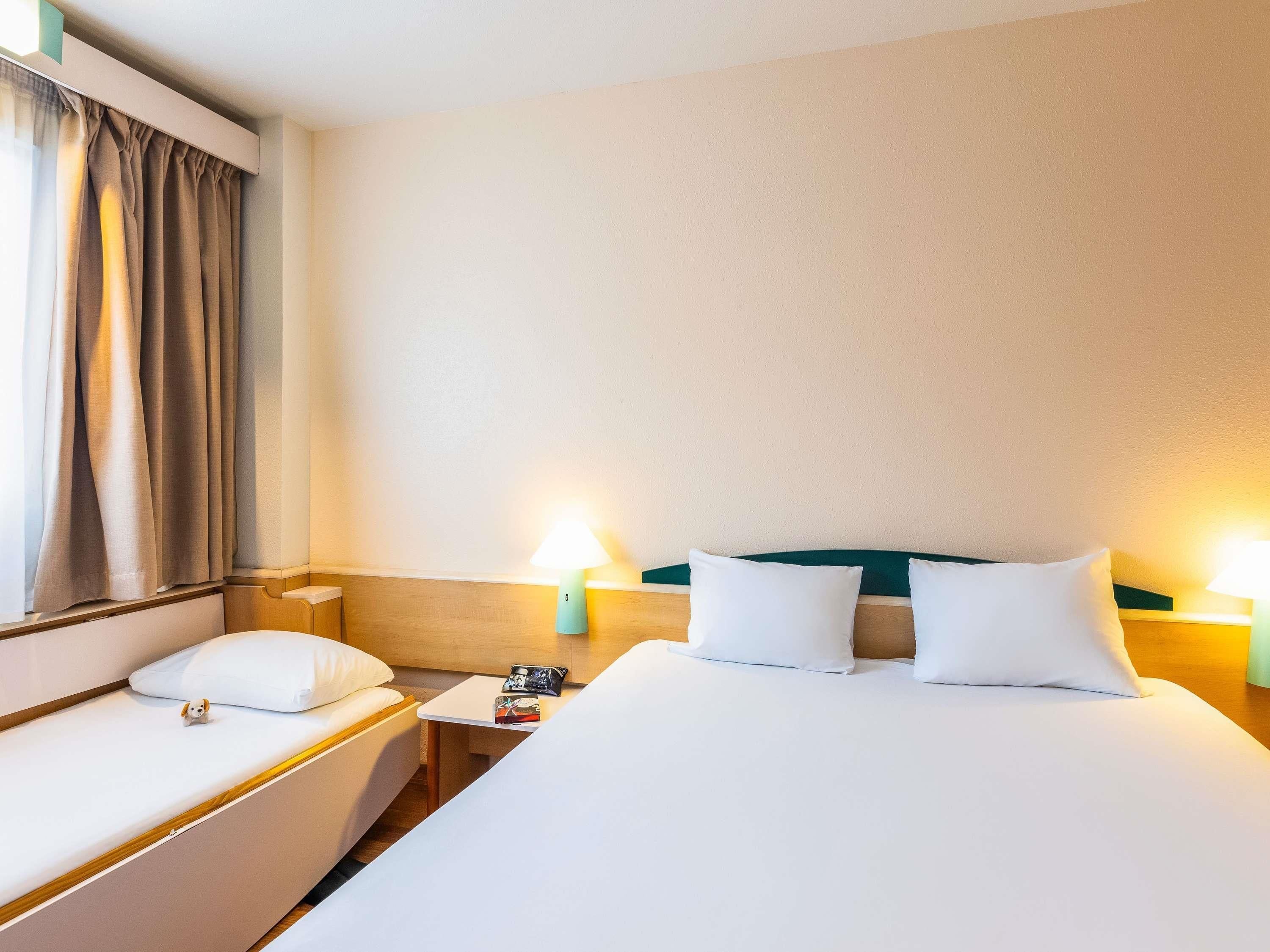 Habitación de hotel con cama, dos mesitas de noche, una lámpara y un ventanal con cortinas.