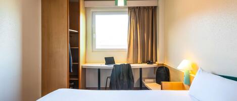 A hotel room with a bed, a desk with a laptop, a chair, a lamp, and a window with curtains.