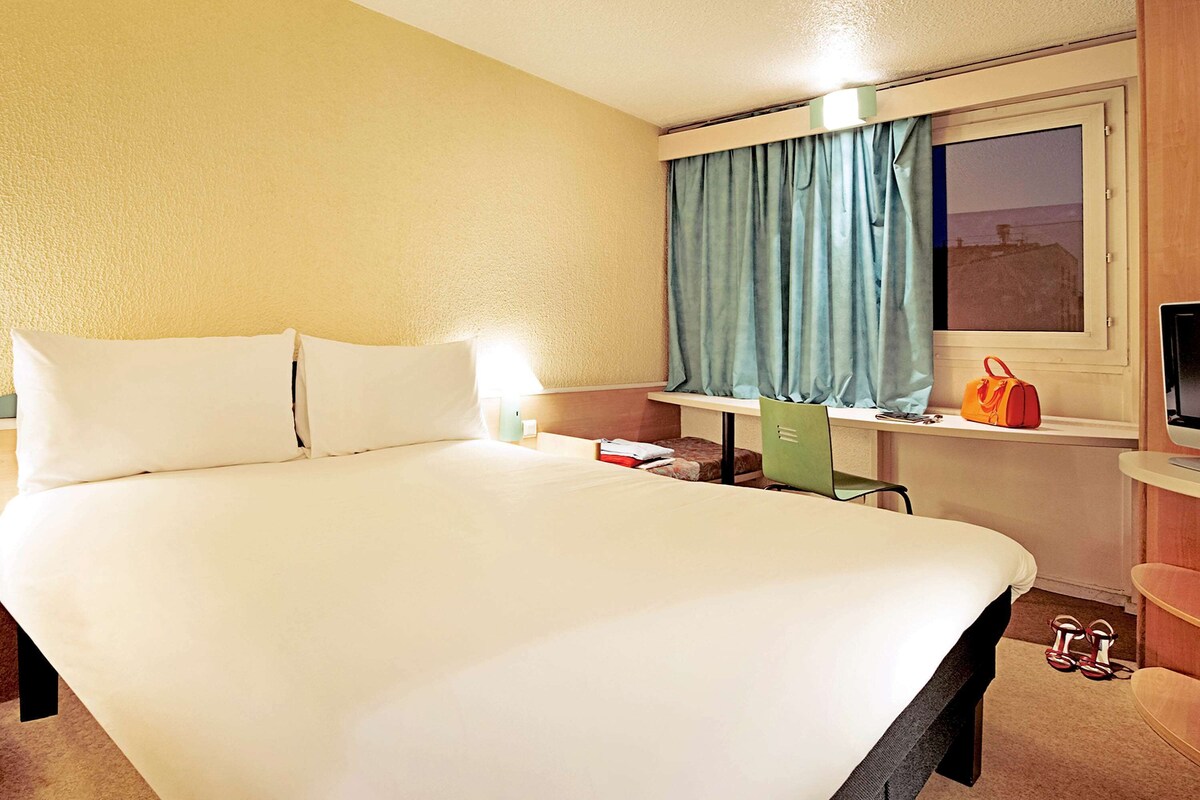 Una habitación de hotel con una cama grande, un escritorio con una silla, una ventana con cortinas, un televisor y un bolso sobre el escritorio.