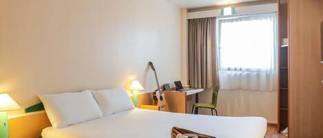 A hotel room with a bed, bedside table, lamp, guitar, desk, chair, and a wardrobe.