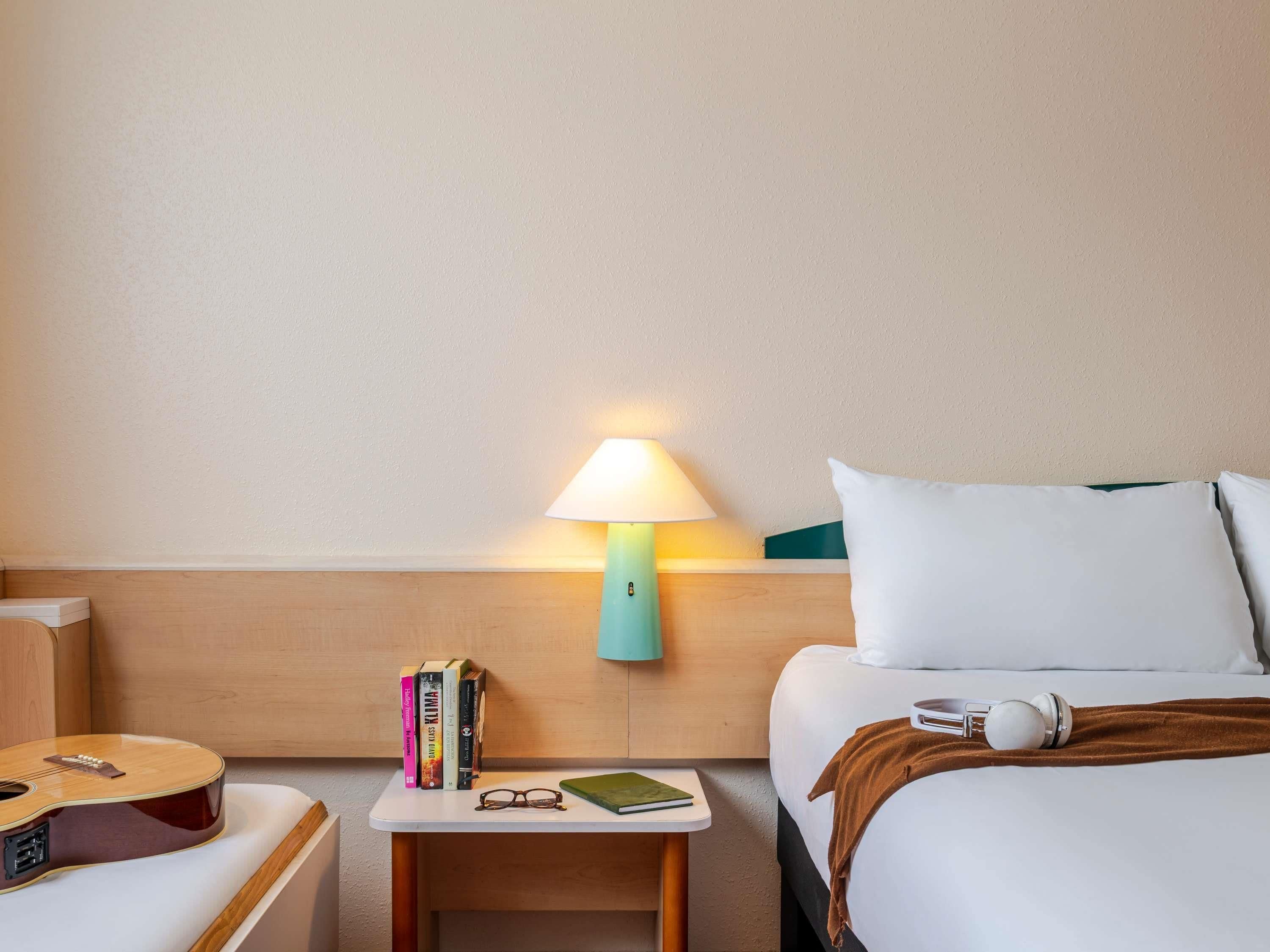 A hotel room with a wooden headboard, a bedside table with a lamp, books, glasses, a guitar, and a bed with pillows and a blanket.