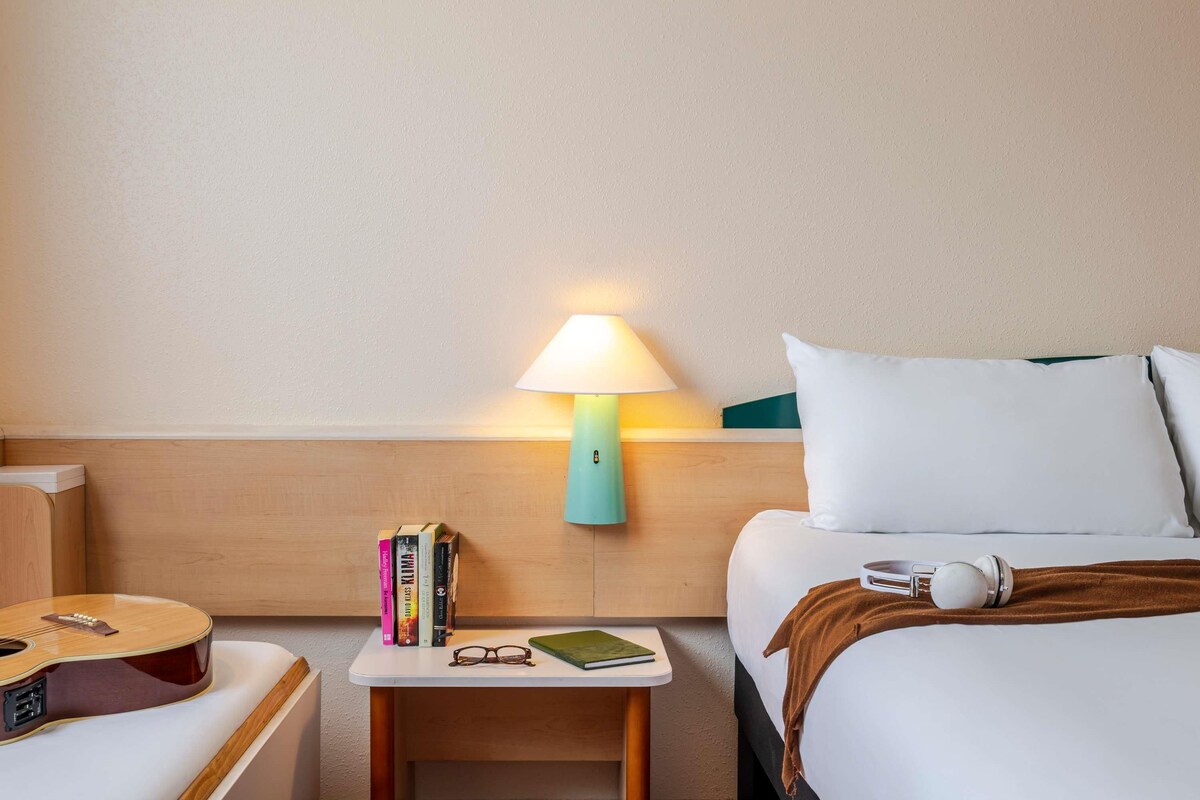 A hotel room with a wooden headboard, a bedside table with a lamp, books, glasses, a guitar, and a bed with pillows and a blanket.