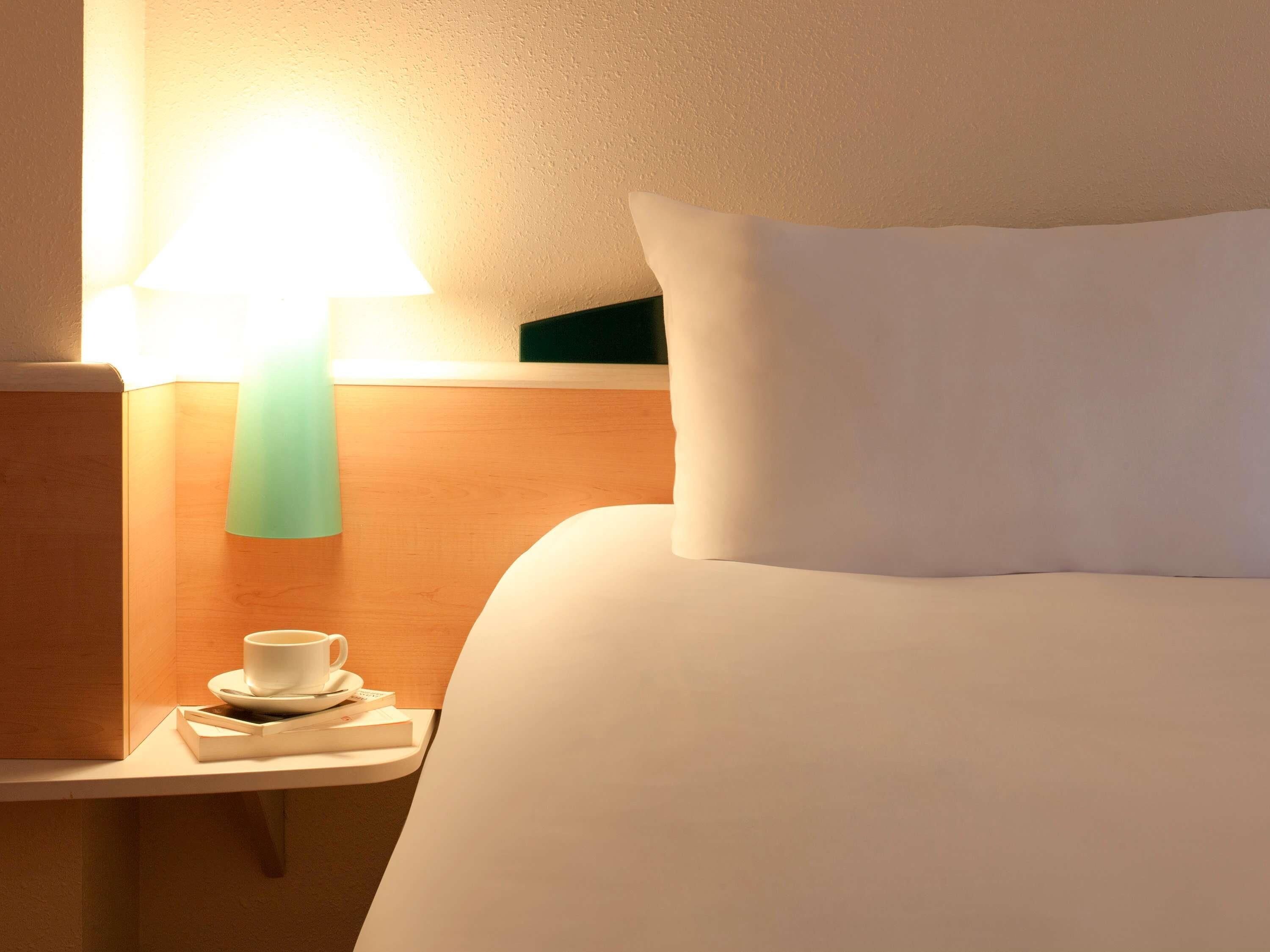 Een hotelkamer met een netjes opgemaakt bed, een nachtkastje met een kop en boek, en een wandlamp.