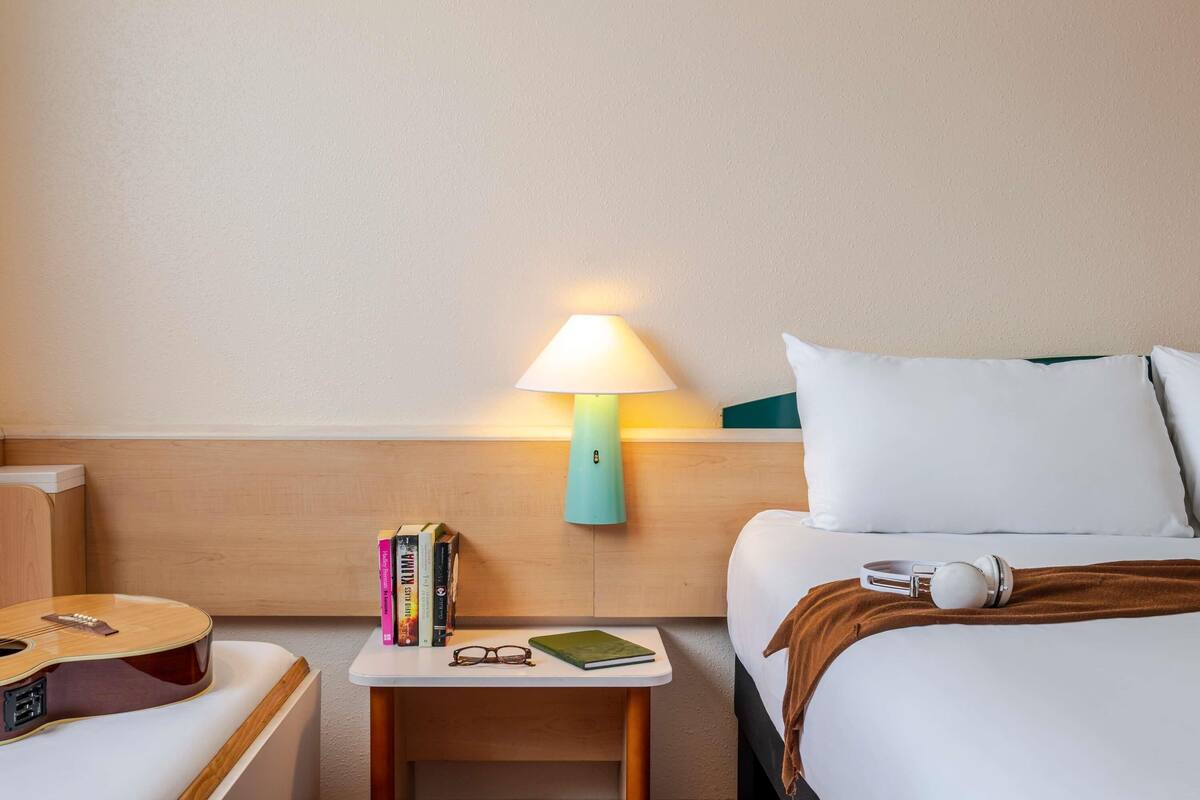 A hotel room with a wooden headboard, a bedside table with a lamp, books, glasses, a guitar, and a bed with pillows and a blanket.