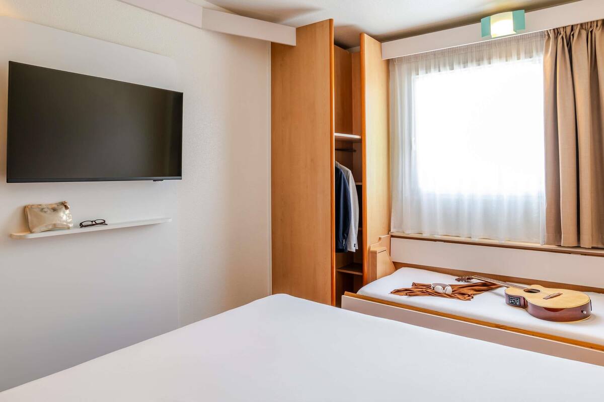A hotel room with a bed, a TV mounted on the wall, a window with curtains, and a wooden wardrobe.
