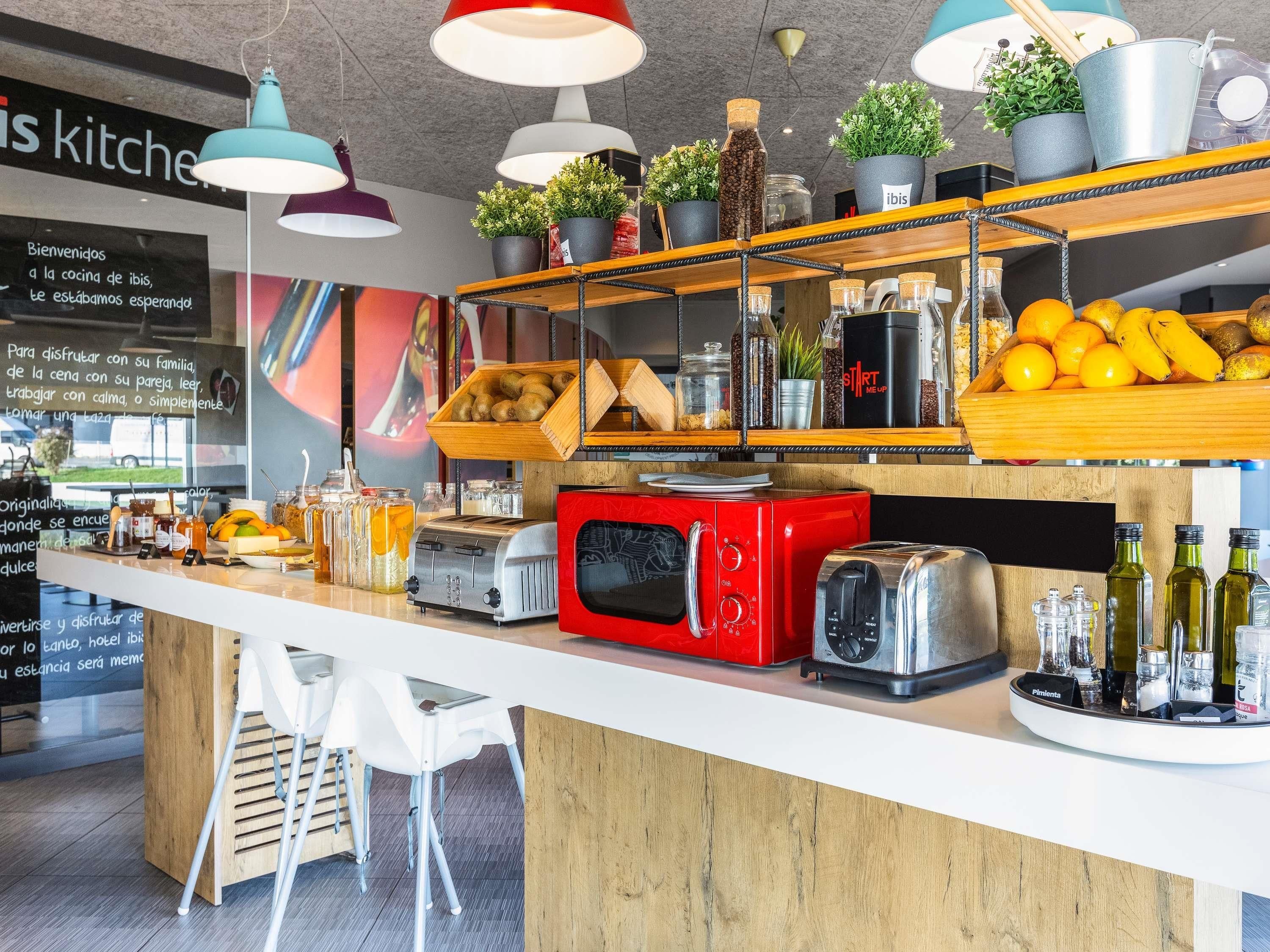 A kitchen area with a wooden counter, various appliances including a red microwave and a toaster, and shelves with fruits and jars.
