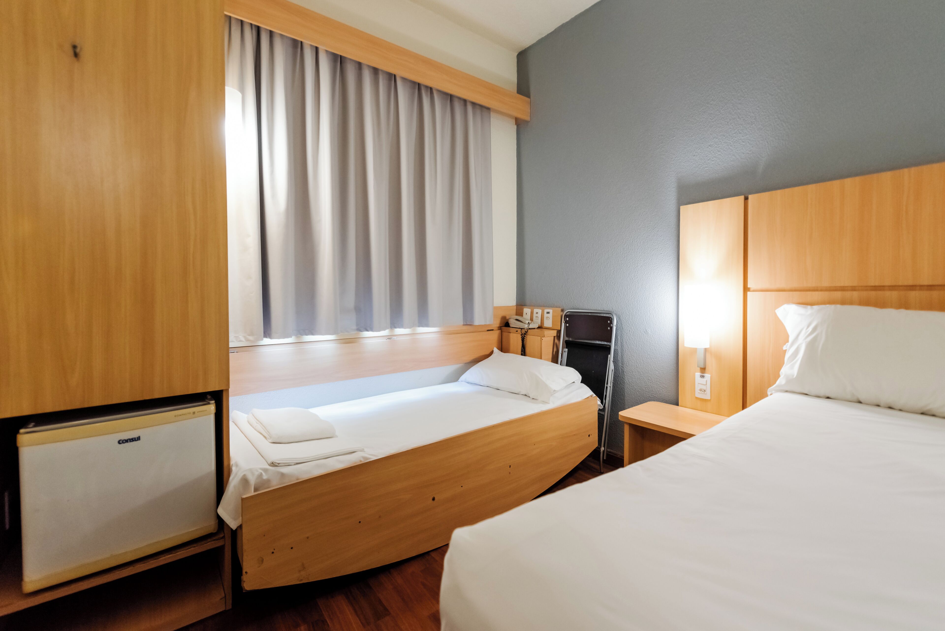 A hotel room with a bed, a desk, a lamp, a mini-fridge, and a window with curtains.