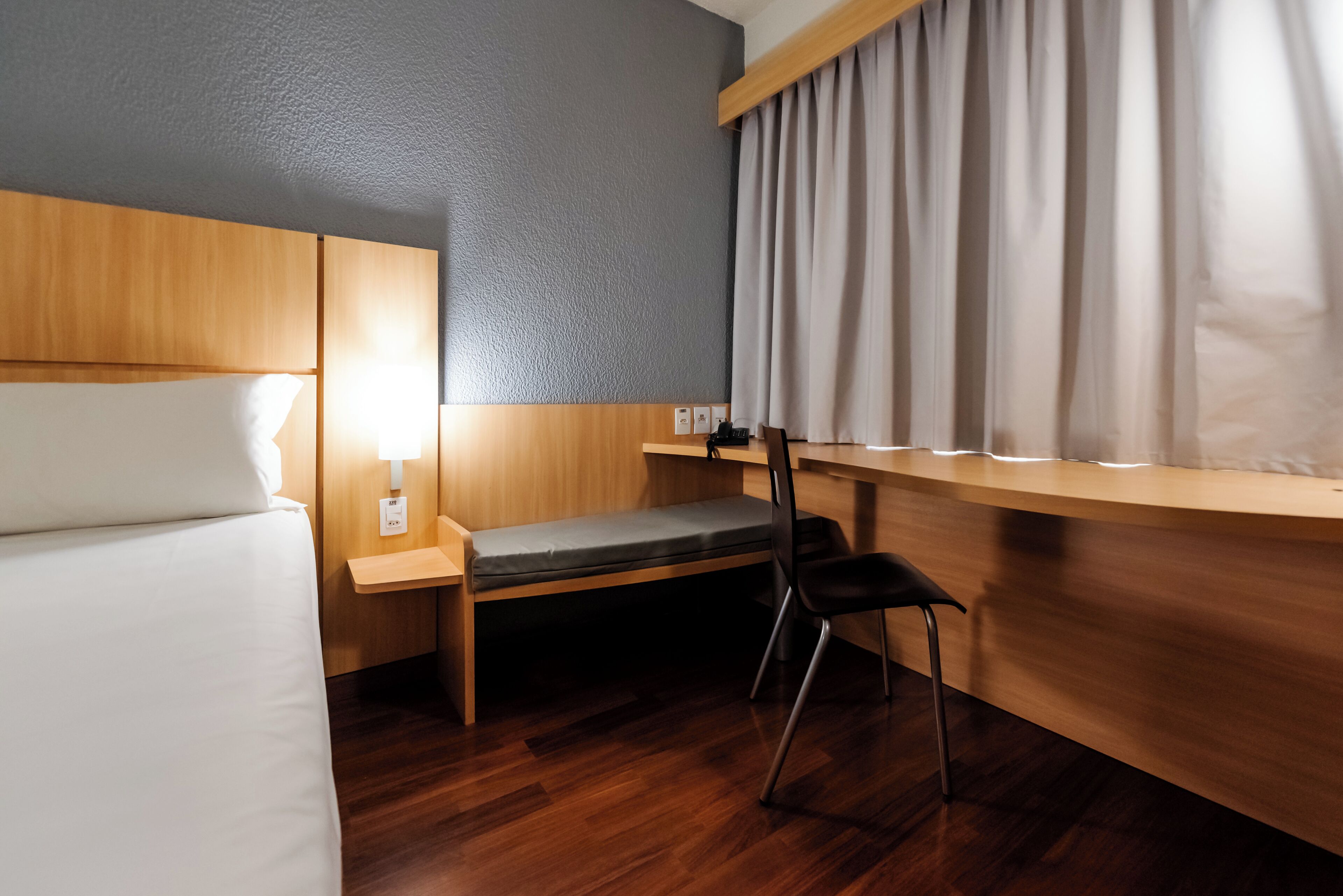A hotel room with a bed, a desk, a chair, a lamp, and a window with curtains.
