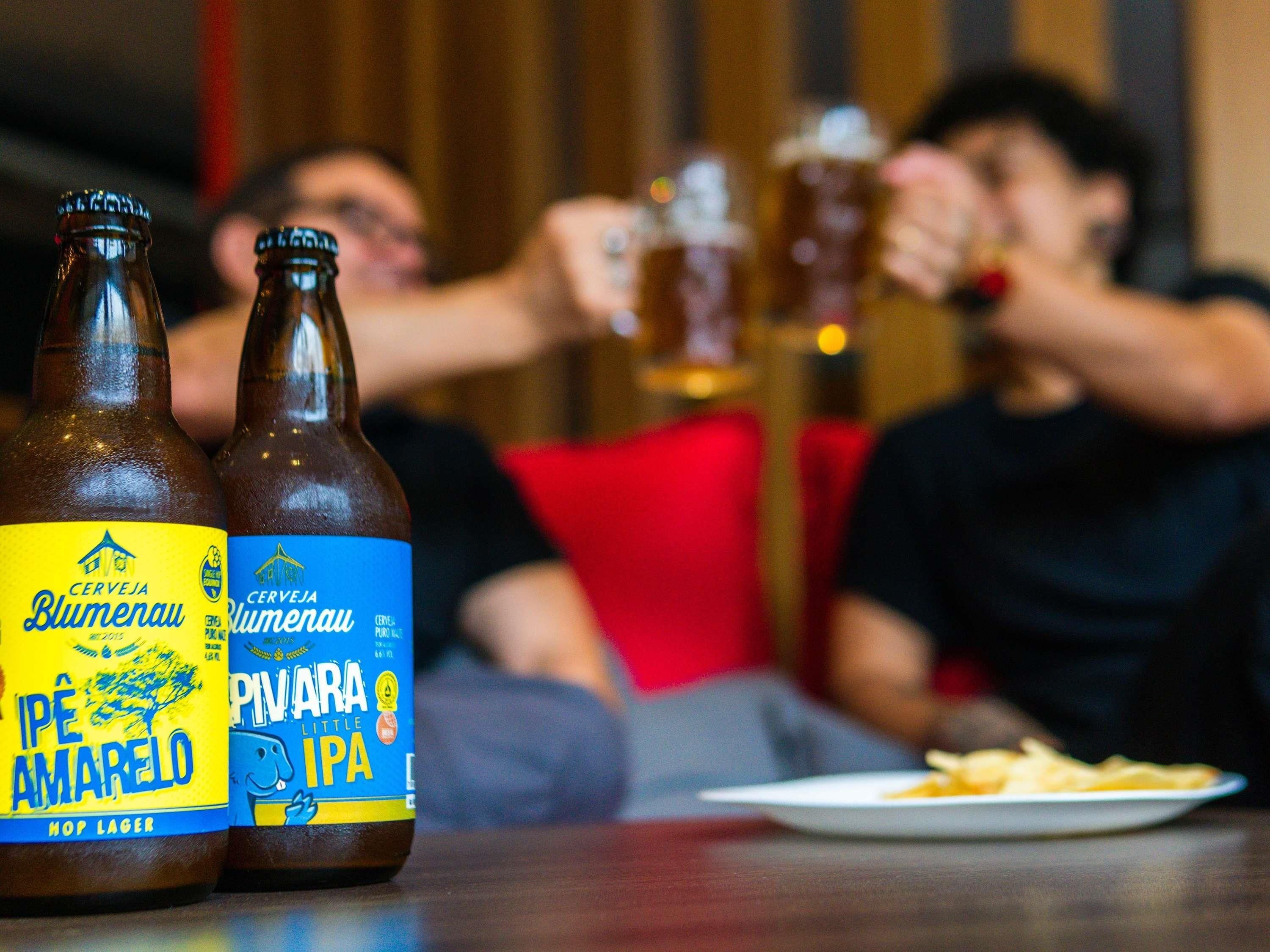 Two bottles of beer on a table with a plate of food and a person in the background.