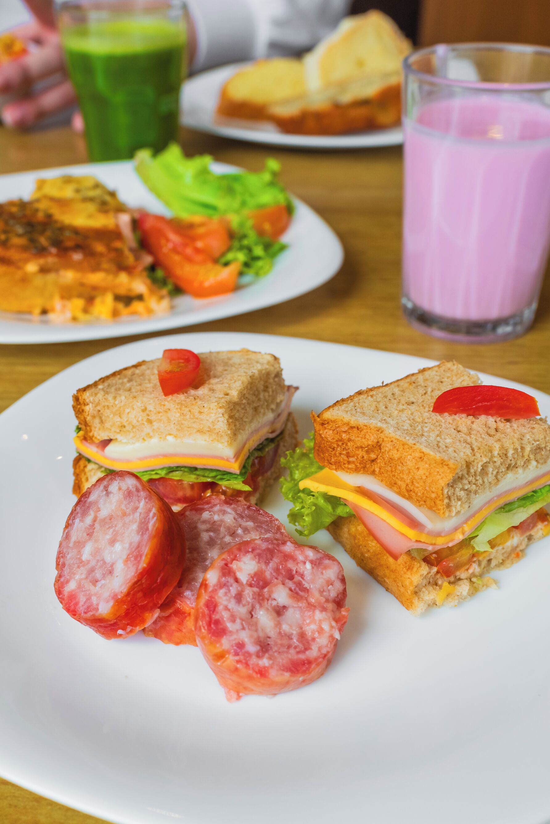 A plate with a sandwich, slices of sausage, and a glass of pink drink on a table.
