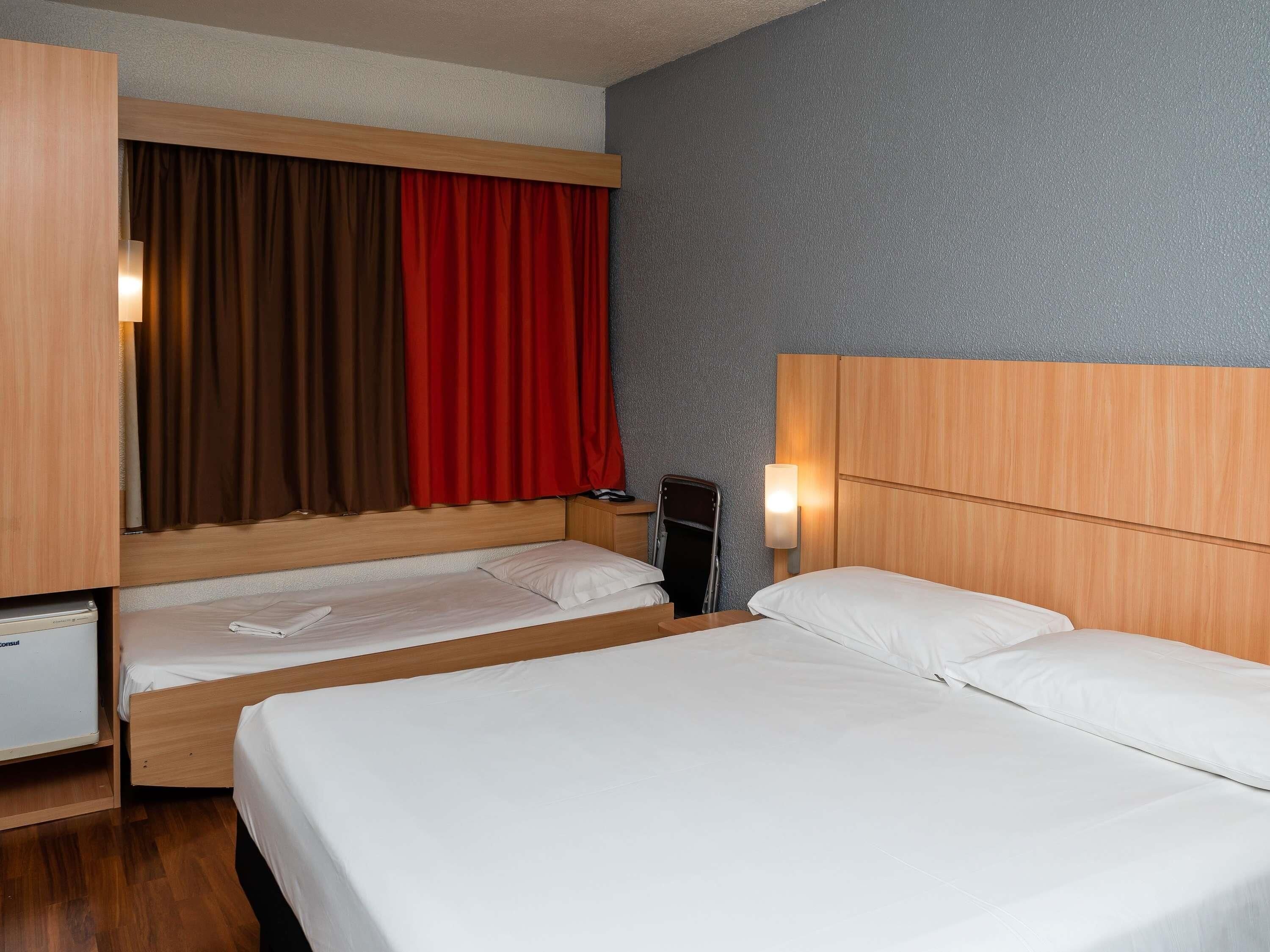 Una habitación de hotel con dos camas, un escritorio, una silla, una mesita y una lámpara fijada a la pared.