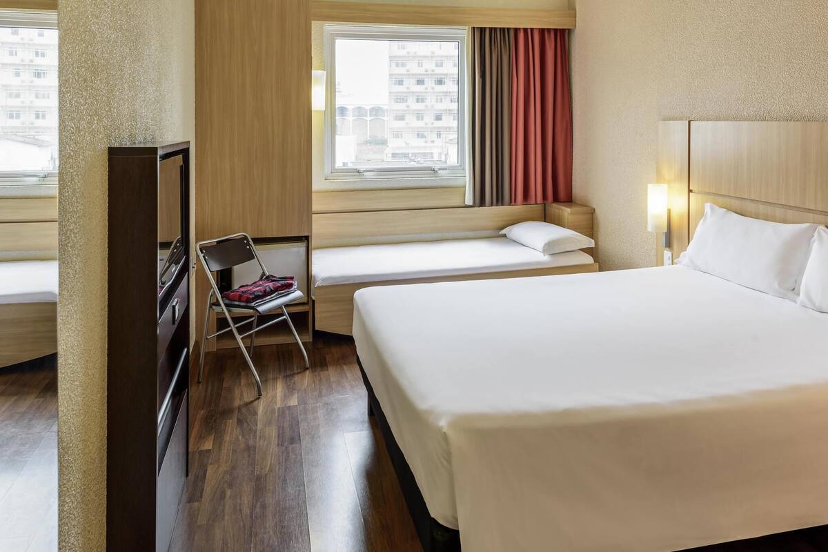 Una habitación de hotel con una cama grande, una cama más pequeña, una silla, un televisor y una ventana con cortinas.