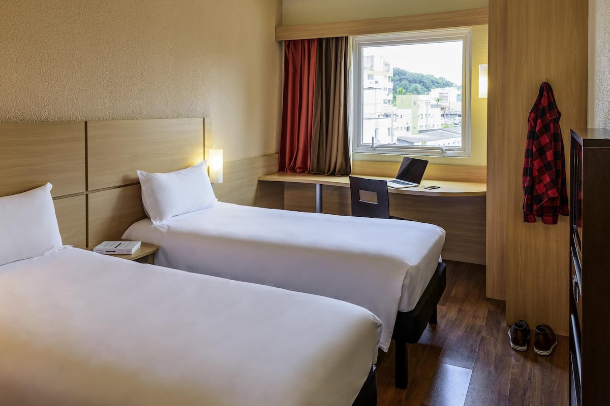 Habitación de hotel con dos camas, un escritorio con una computadora portátil, una ventana con cortinas y un par de zapatos en el suelo.