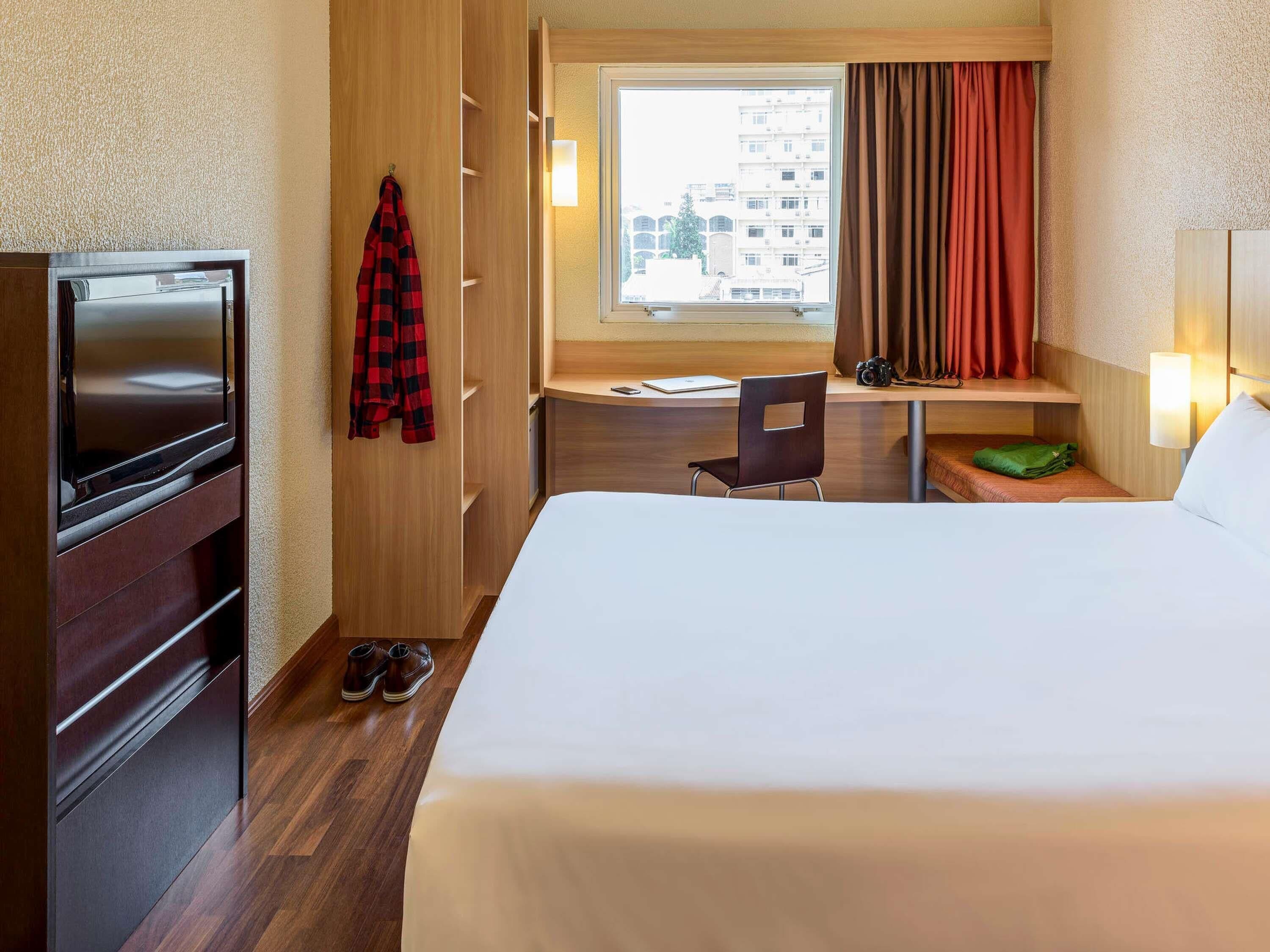 A hotel room with a bed, a desk, a chair, a TV, a coat rack, and a window with curtains.