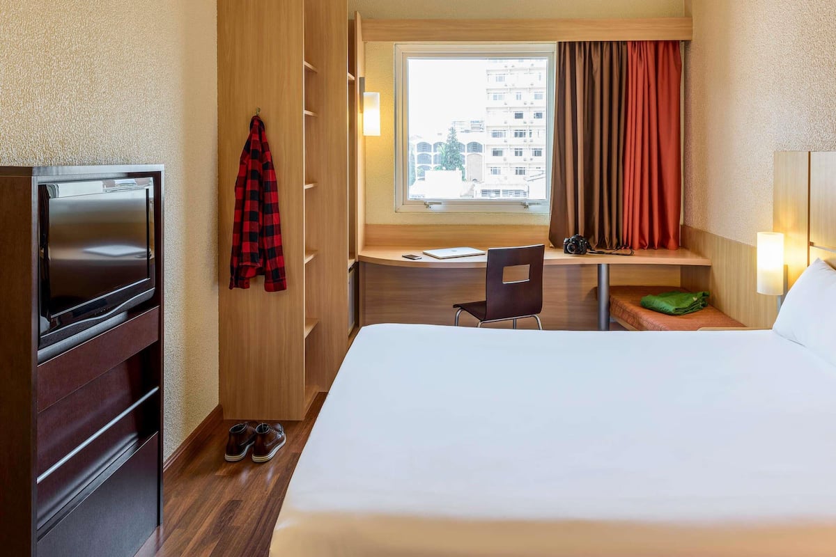 Habitación de hotel con cama, escritorio, silla, televisor, perchero y ventana con cortinas.