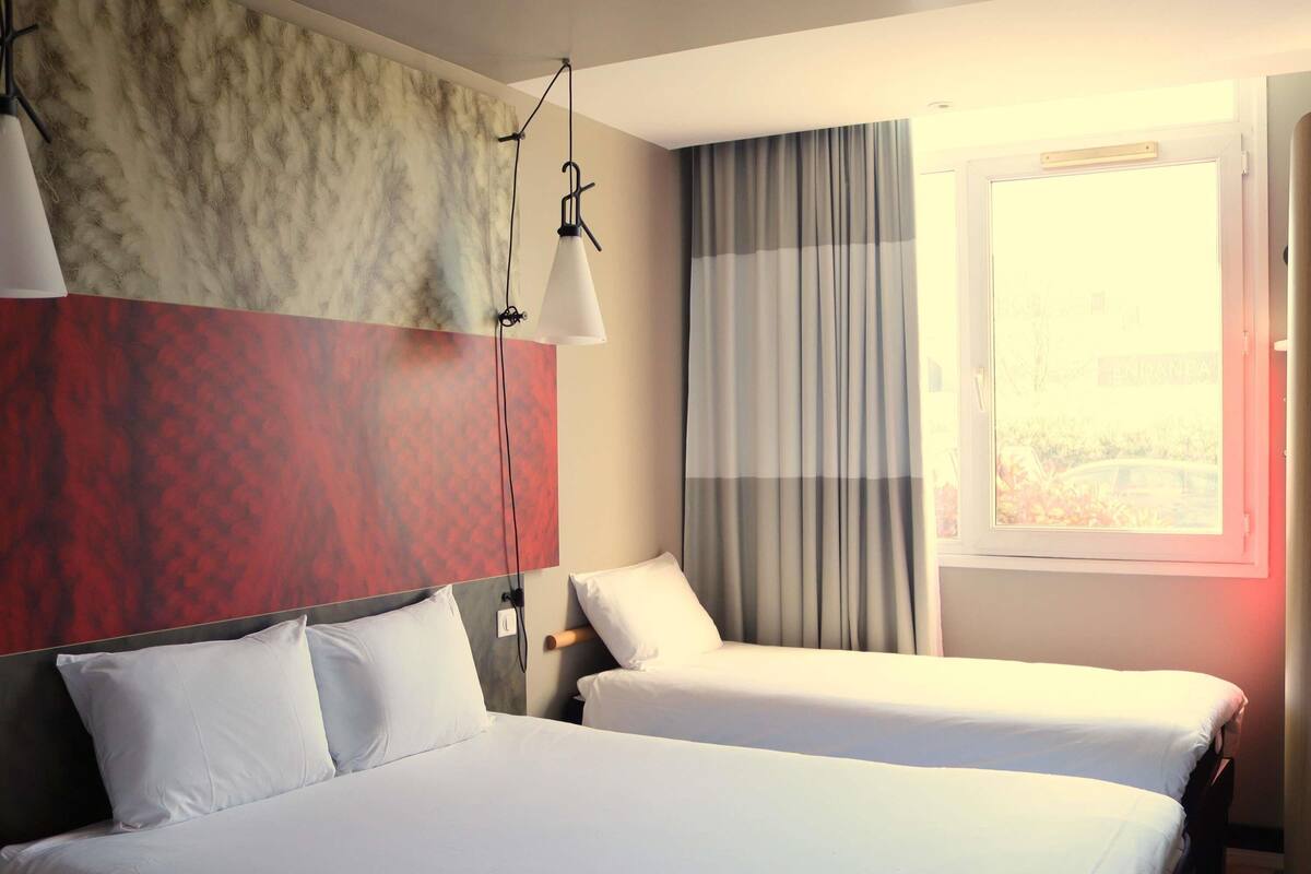 A hotel room with two beds, a large window with curtains, a wall-mounted lamp, and a view of a bridge.