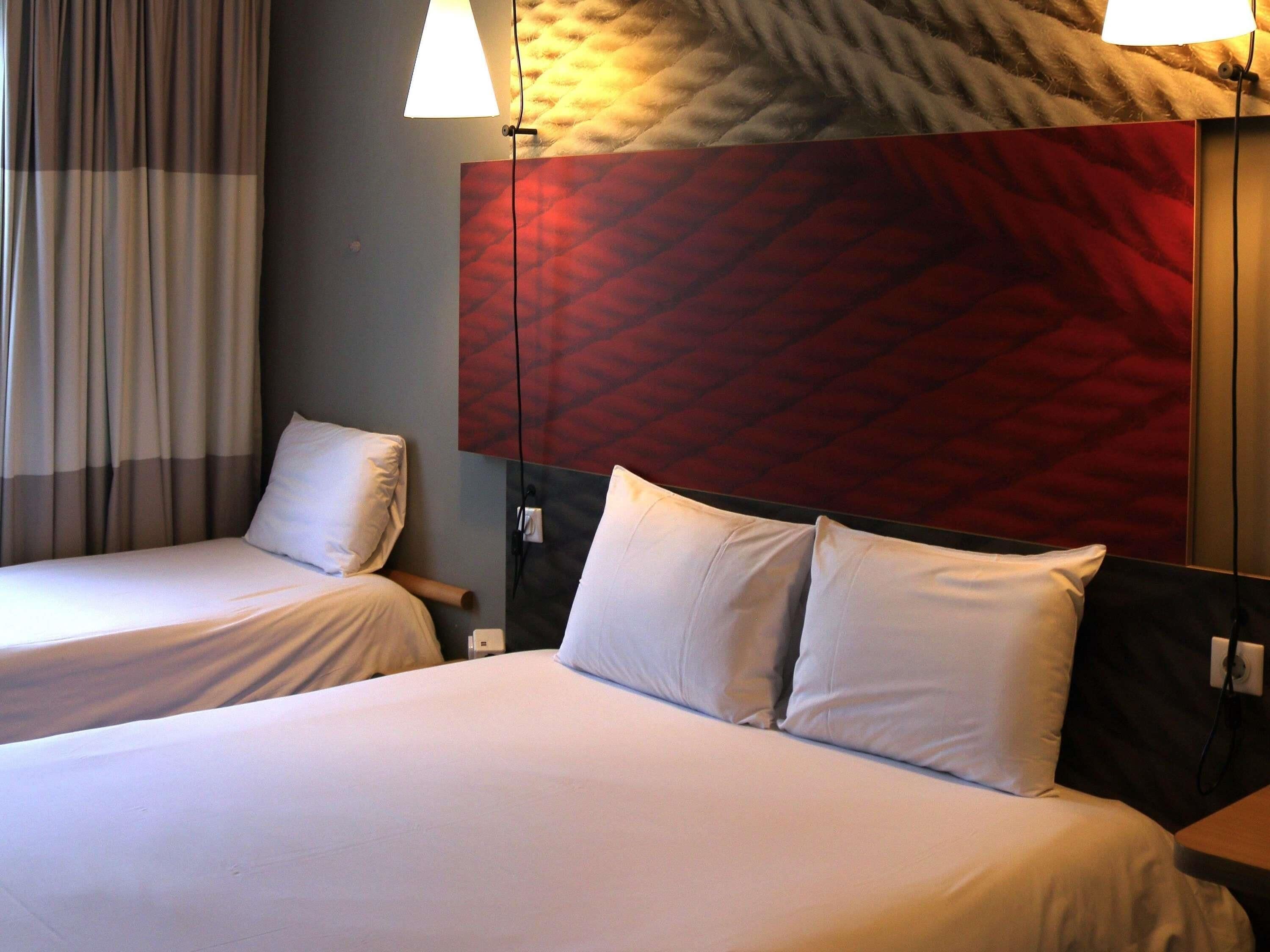Una habitación de hotel con dos camas, un cabecero rojo con textura y una ventana con cortinas.