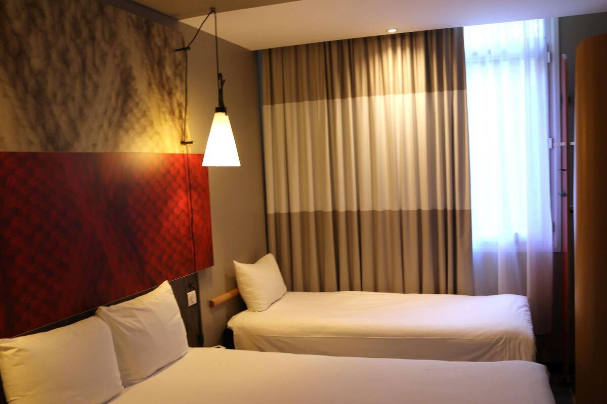 A hotel room with two beds, a red textured wall, a hanging lamp, and a window with curtains.