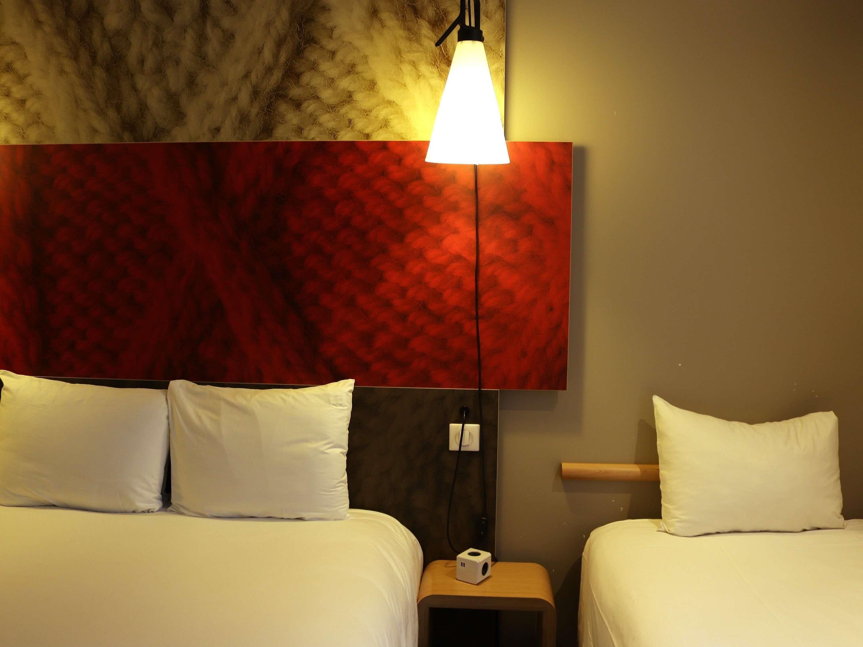 Une chambre d’hôtel avec deux lits, un mur rouge à texture, une lampe blanche et une petite table sur laquelle se trouve un appareil.